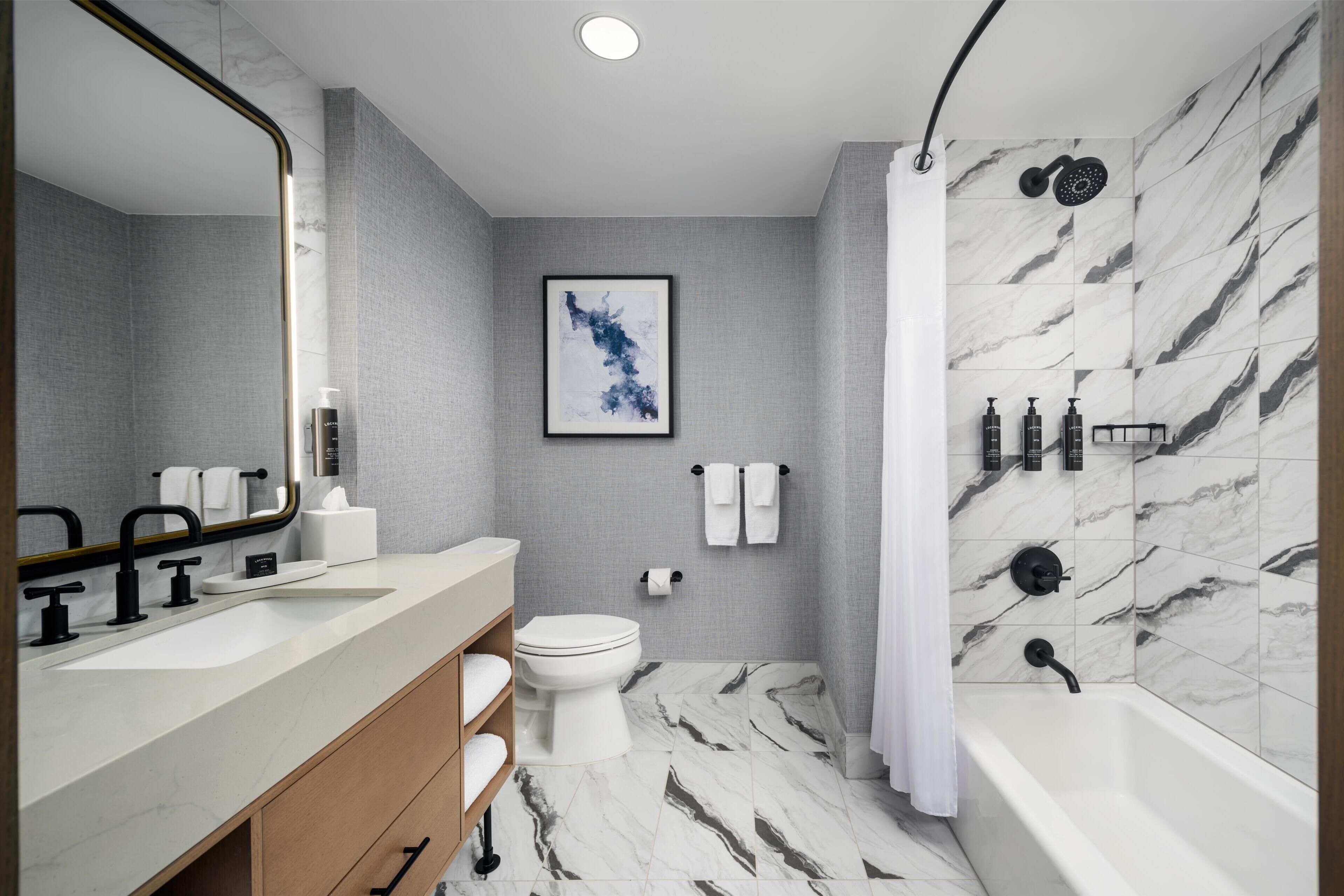 A modern bathroom with a marble floor, a bathtub, a toilet, a sink with a mirror, and a framed artwork on the wall.