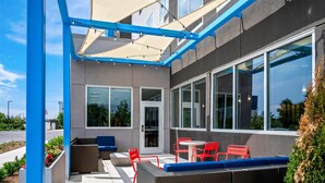 Een terras buiten met rieten meubilair en rode stoelen, grote ramen en een blauw frame.