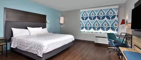 Une chambre d’hôtel avec un grand lit, un bureau avec une chaise, une télévision et une fenêtre ornée d’un motif géométrique.