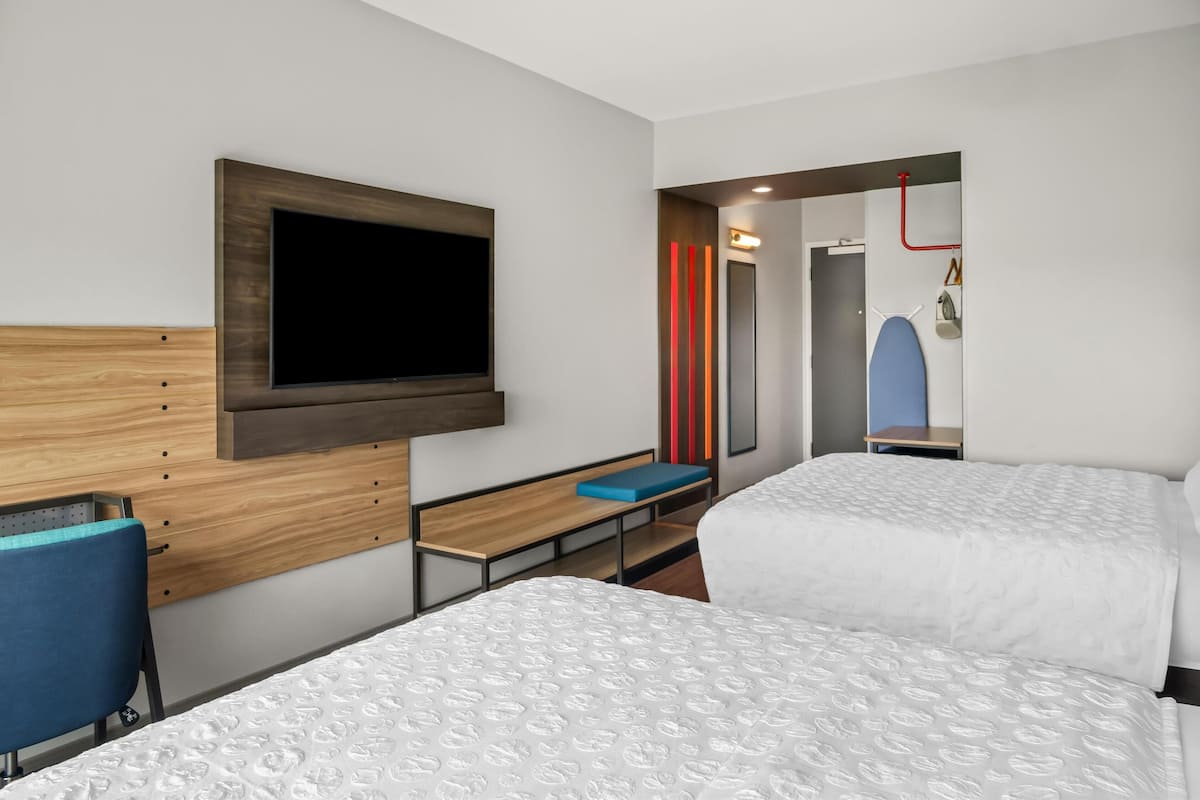 A hotel room with two beds, a TV mounted on the wall, a desk with a chair, and a small seating area.