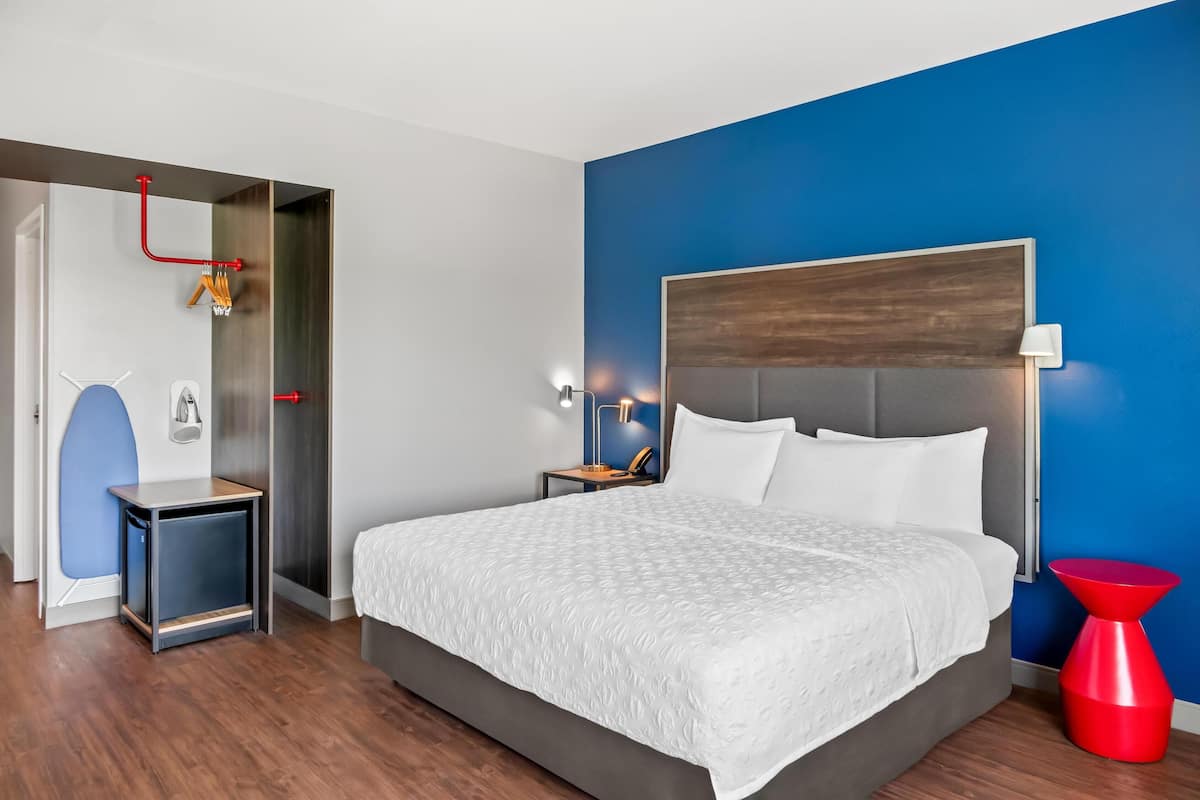 A modern hotel room with a large bed, a nightstand, a red accent chair, and a blue accent wall.