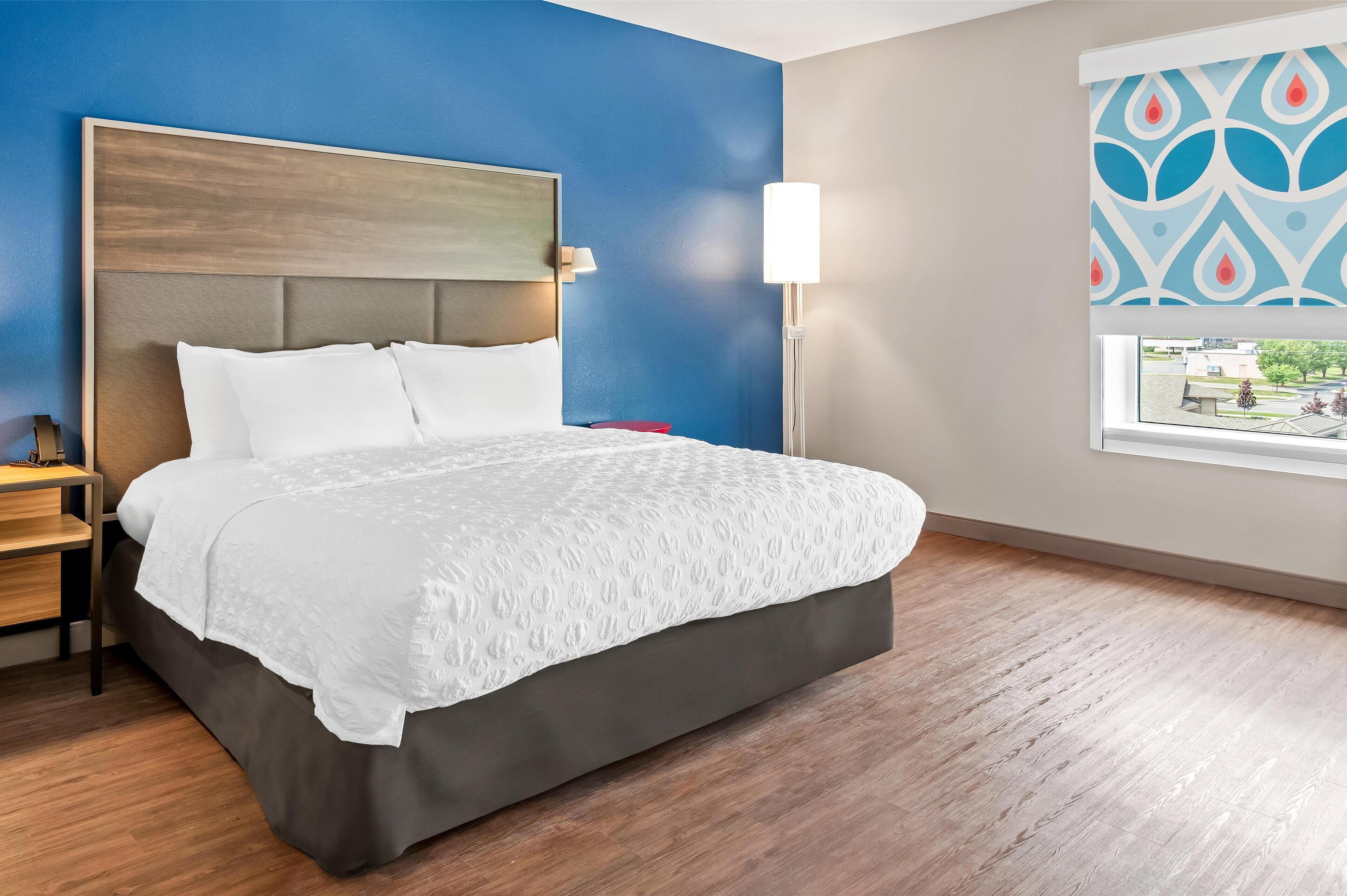 A hotel room with a large bed, a nightstand, a lamp, and a window with a view of the outside.