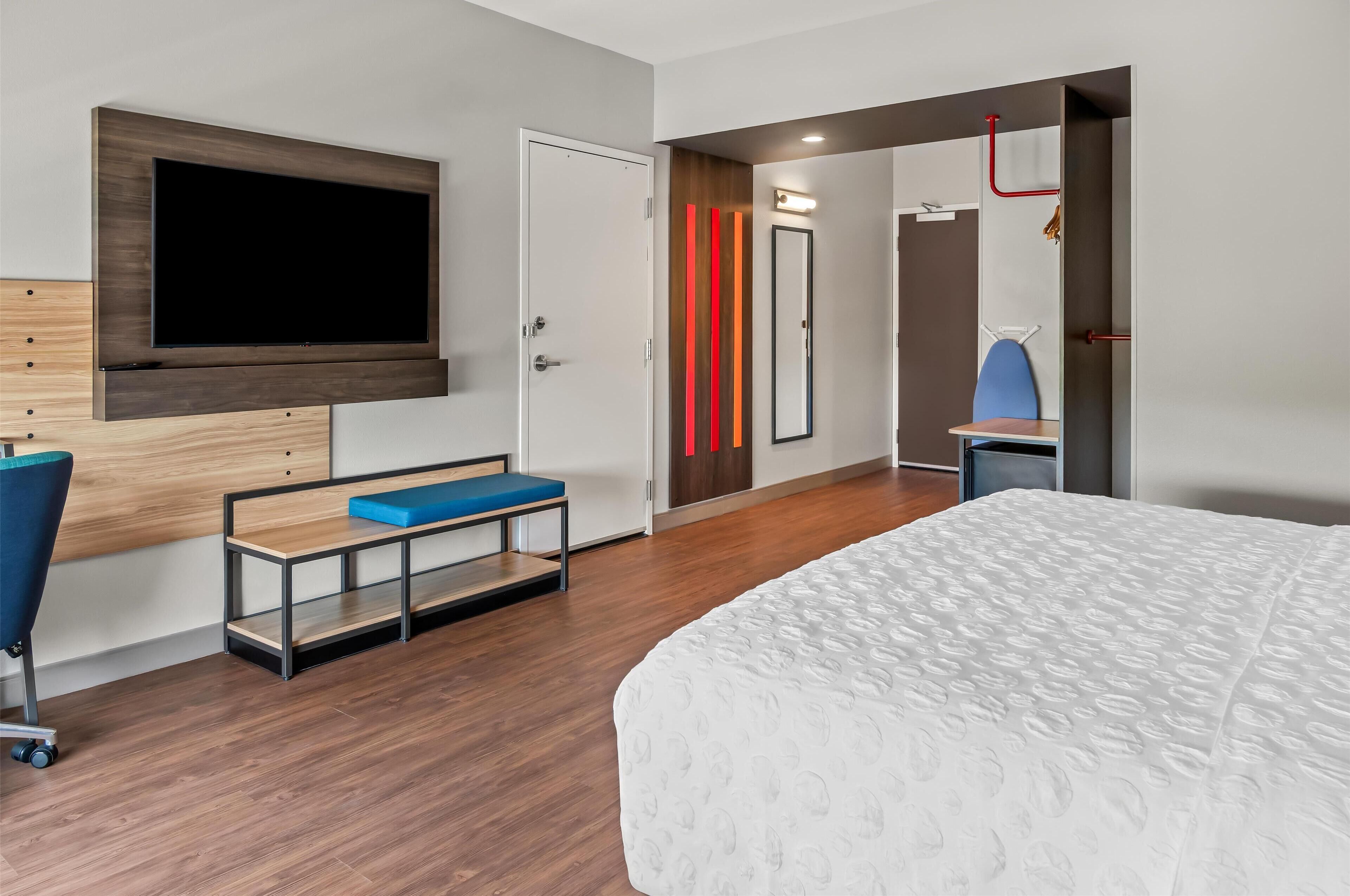 A modern hotel room with a large bed, a TV mounted on the wall, a desk with a chair, and a small seating area.