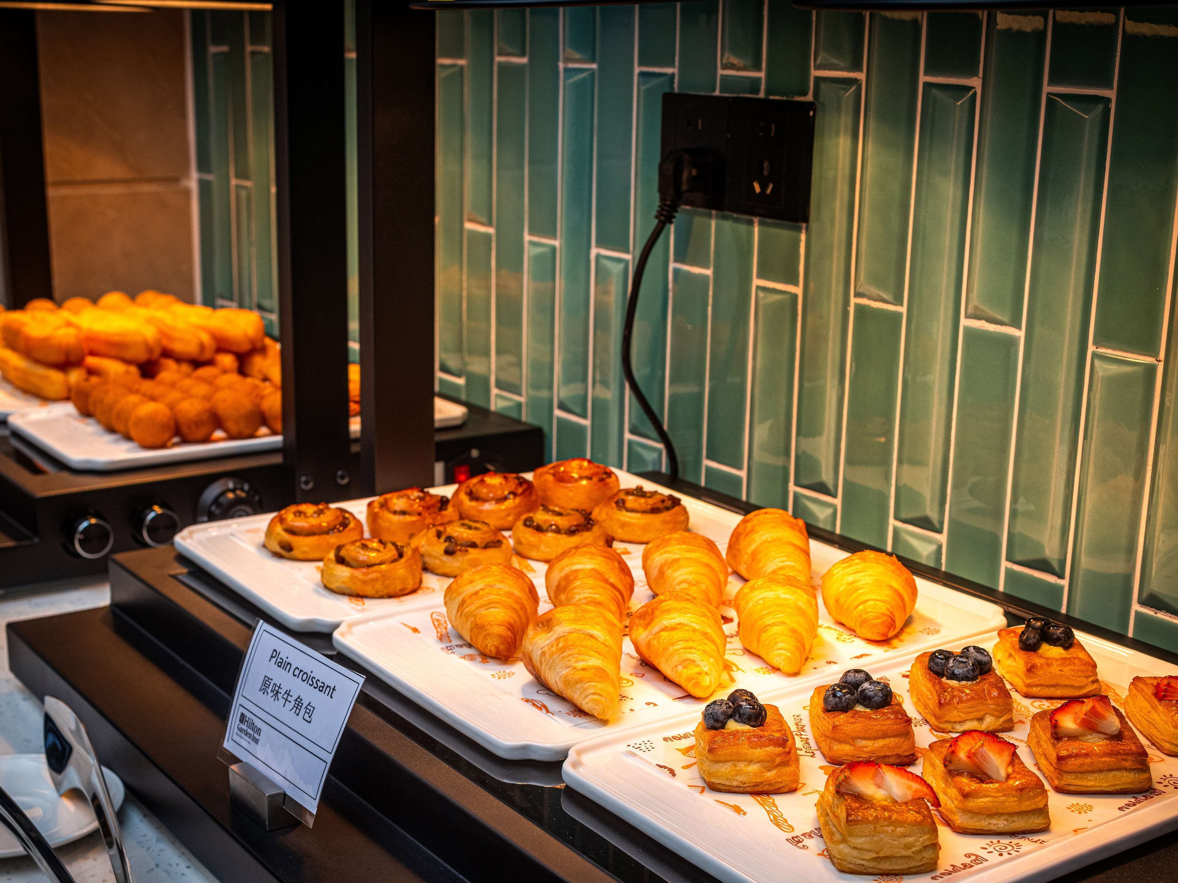 Una exhibición de pasteles que incluye croissants y rollos de canela, con un letrero que indica 'Croissant simple' y 'Wrap original de carne deshebrada'.