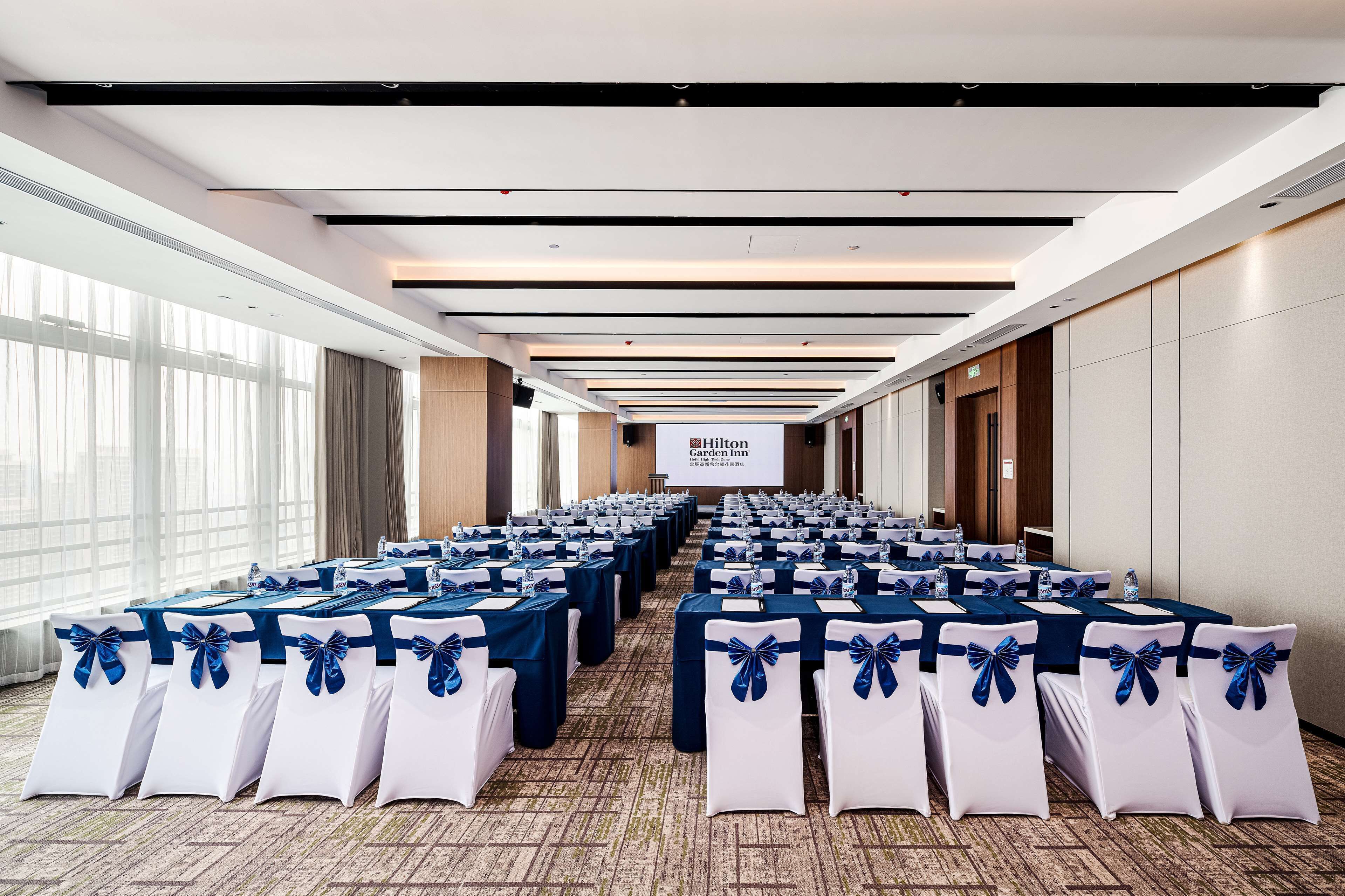 Une salle de conférence avec des rangées de tables et de chaises, un grand écran affichant le nom « Hilton Garden Inn », et de grandes fenêtres sur un des côtés.