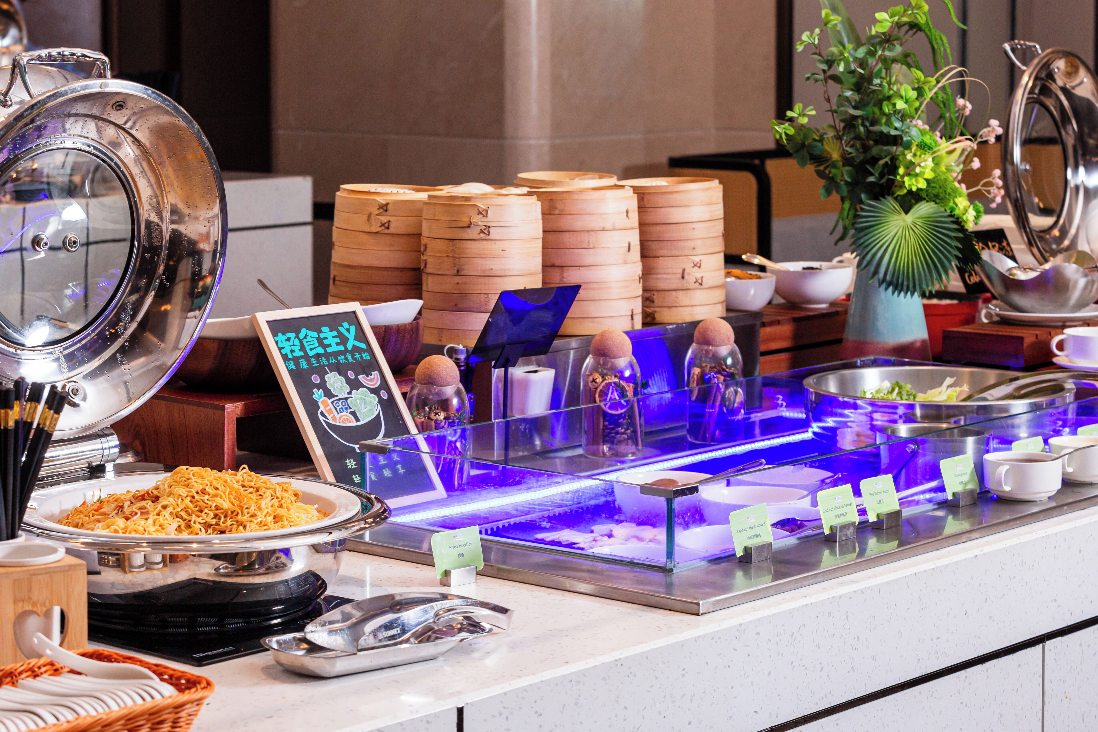 Un buffet proposant une variété de plats, notamment un grand saladier de nouilles, plusieurs cuiseurs à vapeur en bois et une vitrine réfrigérée où les plats sont présentés avec leurs étiquettes.