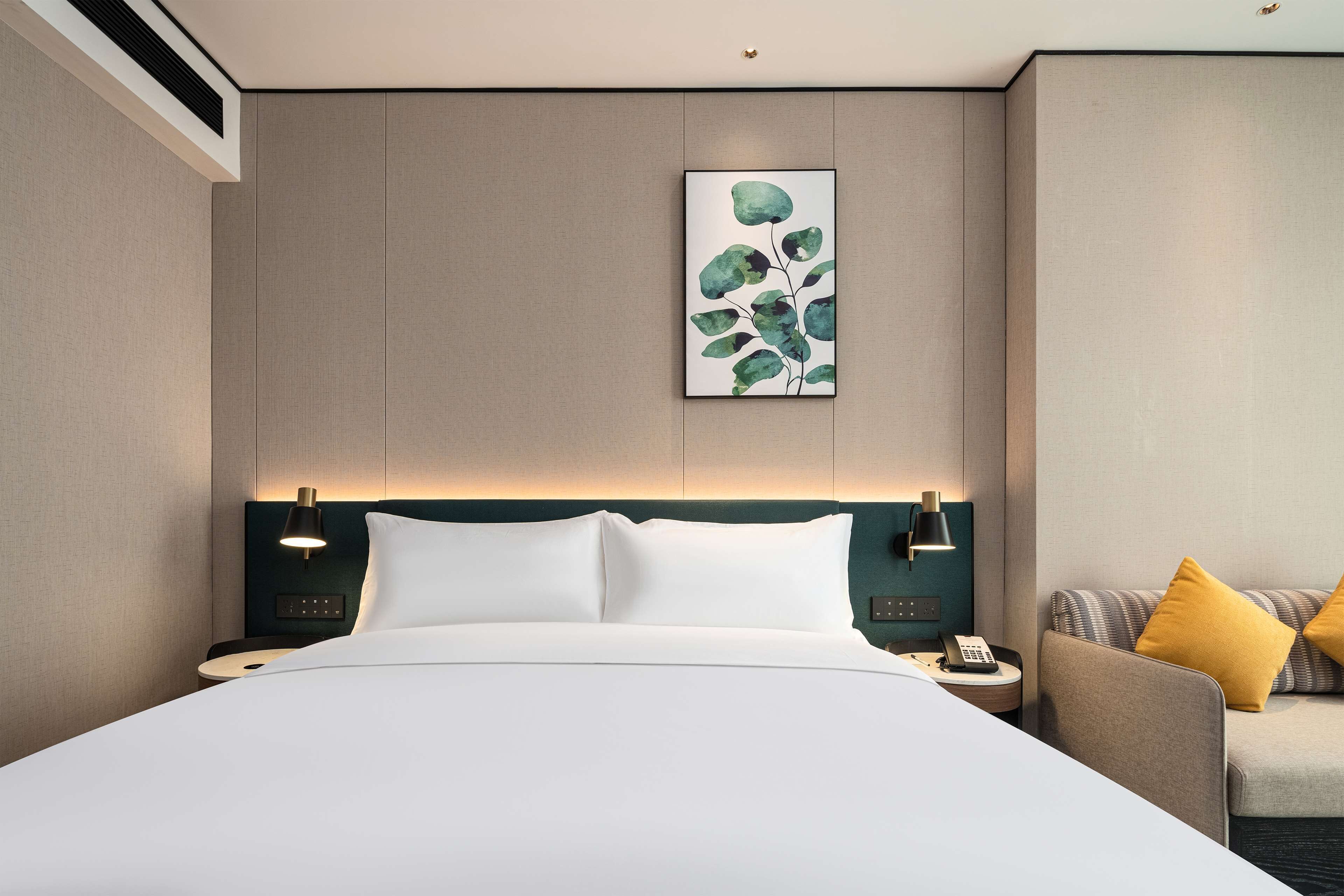 Une chambre d’hôtel moderne avec un grand lit, une table de chevet avec une lampe, une petite table avec un téléphone et un canapé avec des coussins.