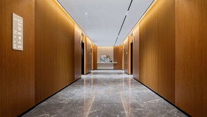 Un couloir moderne avec des lambris en bois et un sol en marbre, menant à un espace d’ascenseur où un panneau indique les niveaux des étages.