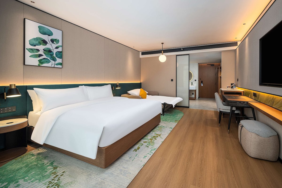 A modern hotel room with a large bed, a desk with a chair, a small seating area, and a bathroom visible through a glass partition.