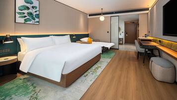 A modern hotel room with a large bed, a desk with a chair, a small seating area, and a bathroom visible through a glass partition.