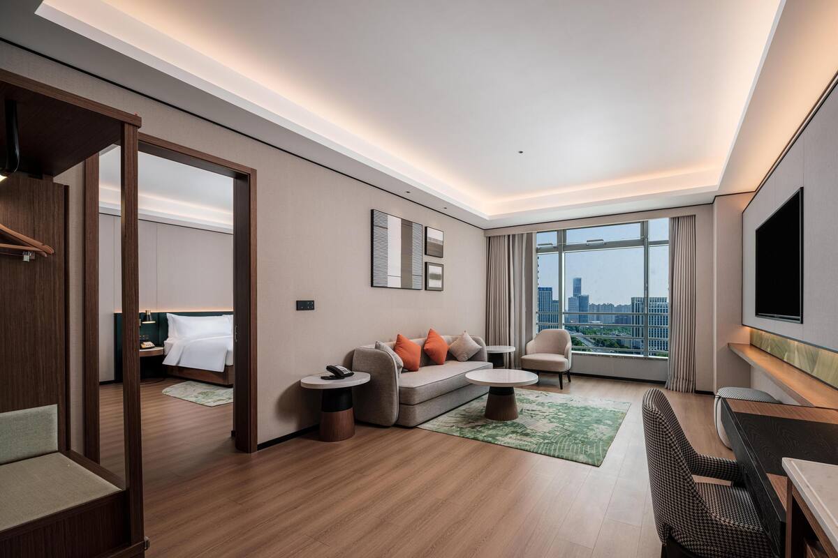 A modern hotel room with a wooden floor, a sofa, a small table, a chair, a TV, and a large window with a city view.