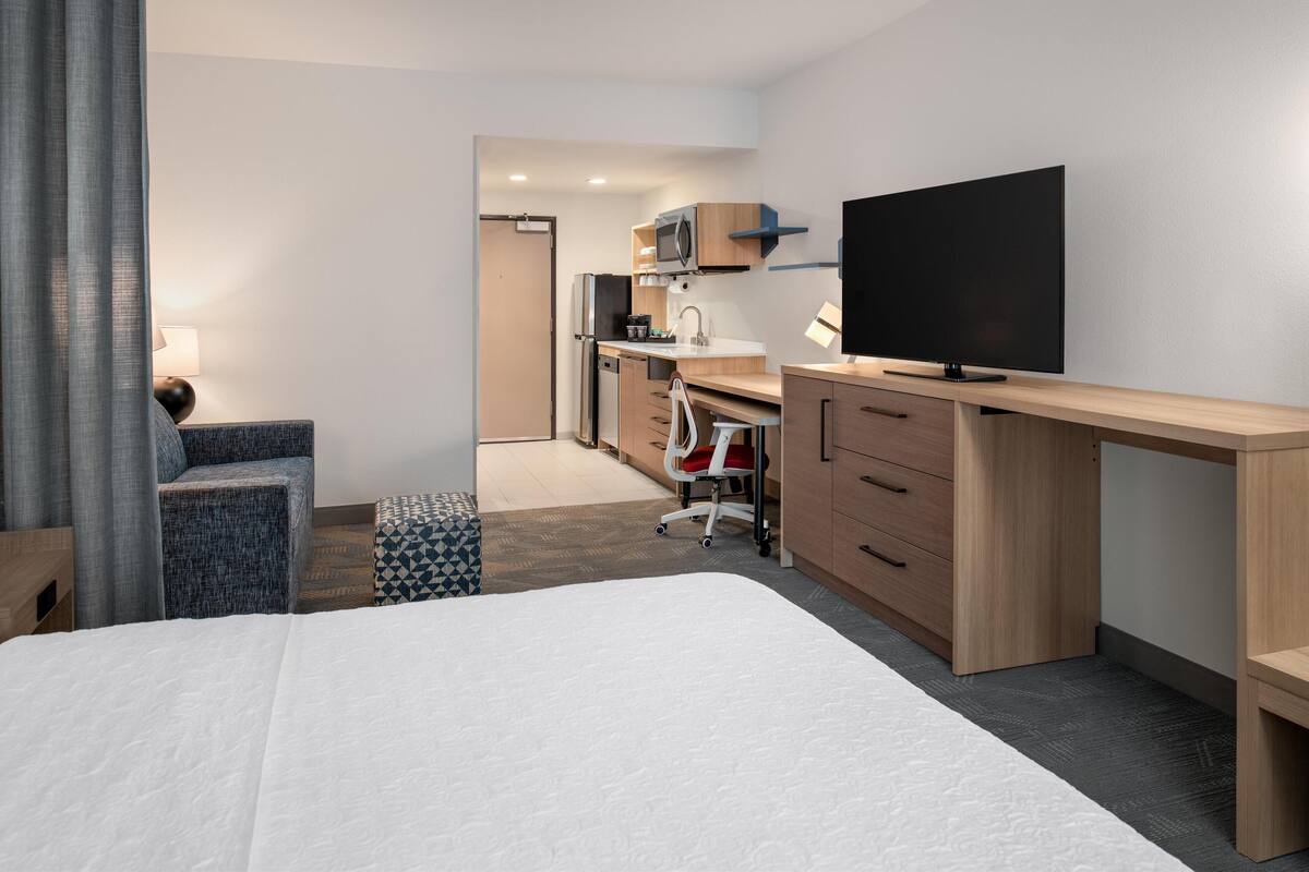 A hotel room with a bed, a desk, a TV, a kitchenette, and a chair.