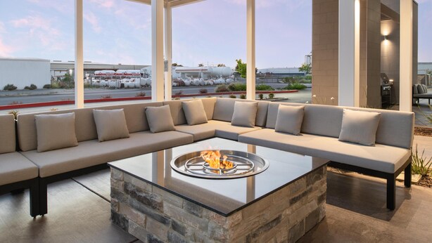 A modern outdoor seating area with a glass-top fire pit, a stone accent wall, and a view of an industrial area. - Home2 Suites By Hilton Woodland Sacramento (Woodland)