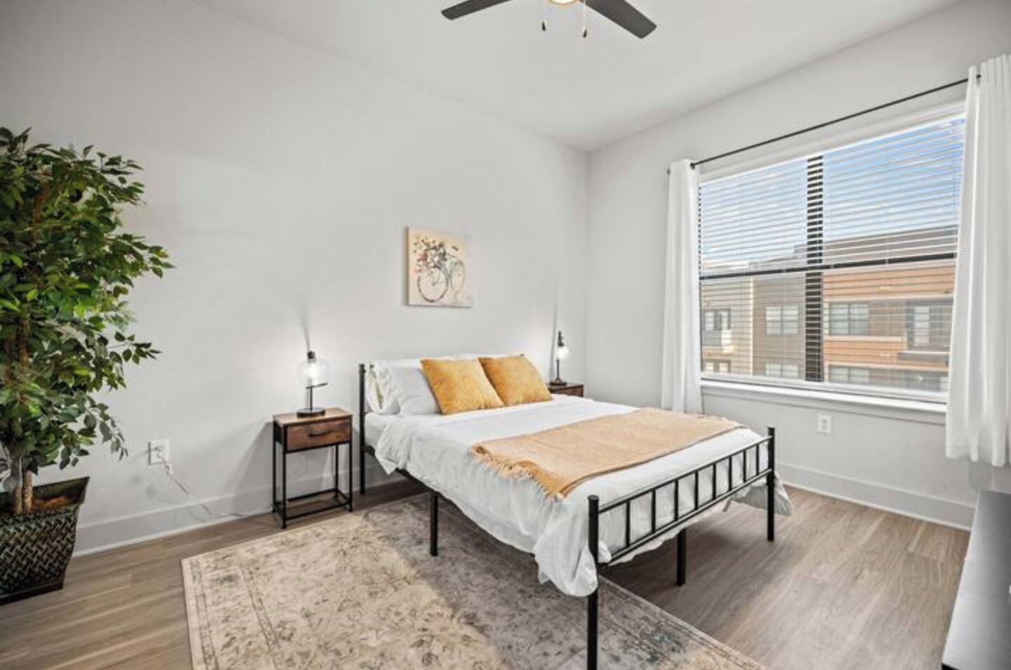 A bedroom with a bed, bedside tables, a floor lamp, a potted plant, and a window with blinds.