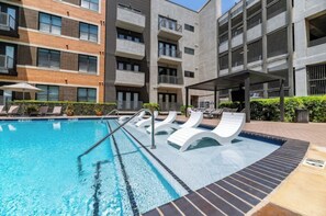 Una piscina con sillas de descanso y una zona de estar cubierta, rodeada de edificios de varios pisos con balcones.