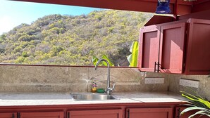 Una cocina roja con un lavamanos y armarios, una ventana con vistas a una colina, y un farol azul colgando arriba.
