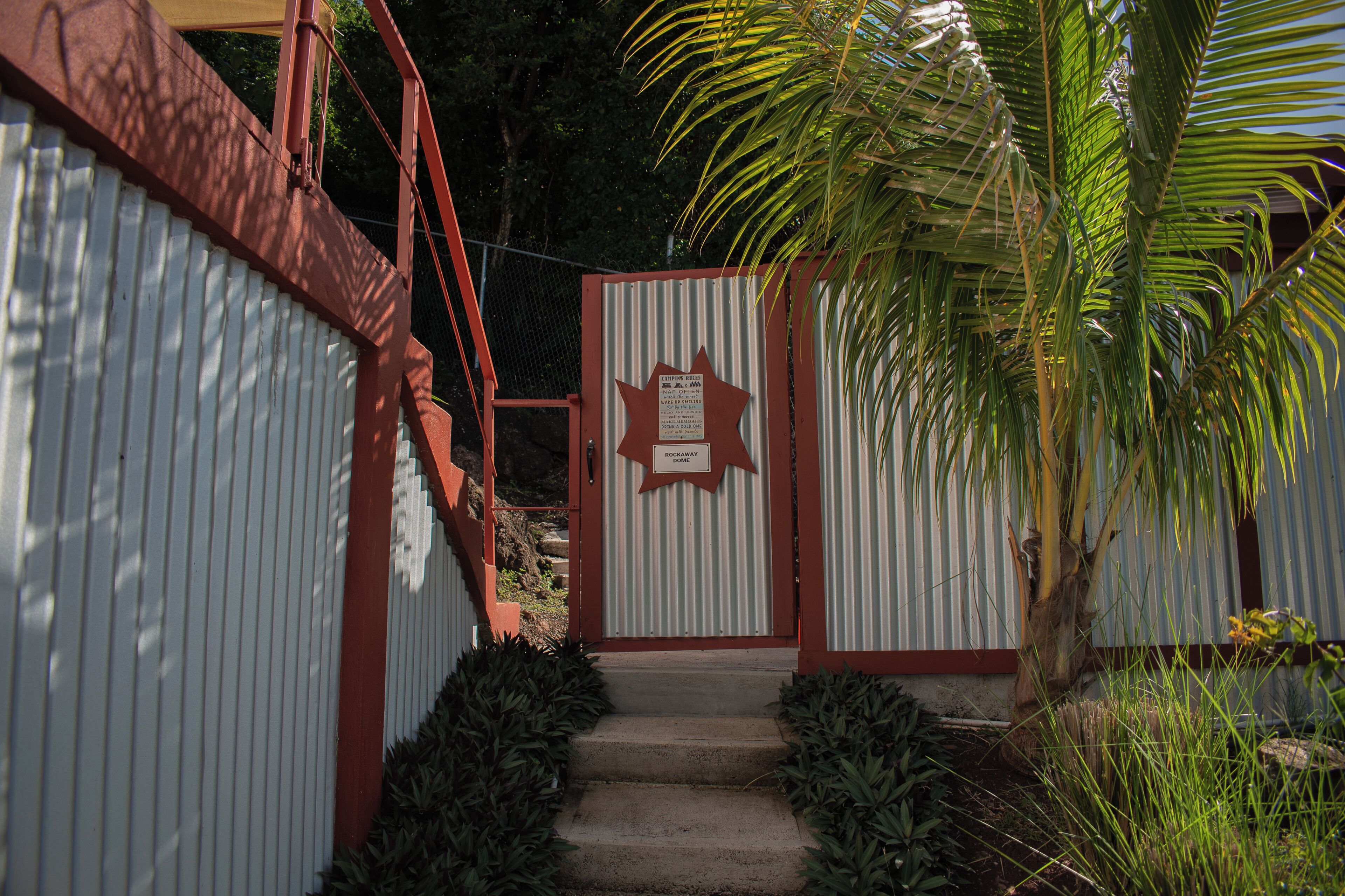 Una entrada con portón, un letrero con forma de estrella, una escalera de metal y una palmera.