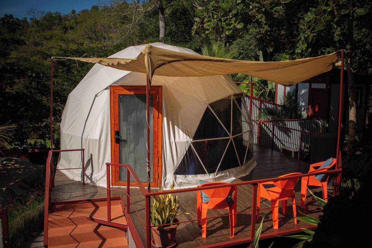 Une tente en forme de dôme avec une porte en bois, une terrasse couverte dotée de rampes rouges et de chaises orange.