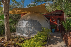 Una estructura de domo geodésico con zona sombreada, rodeada de vegetación tropical y un edificio rojo al fondo.
