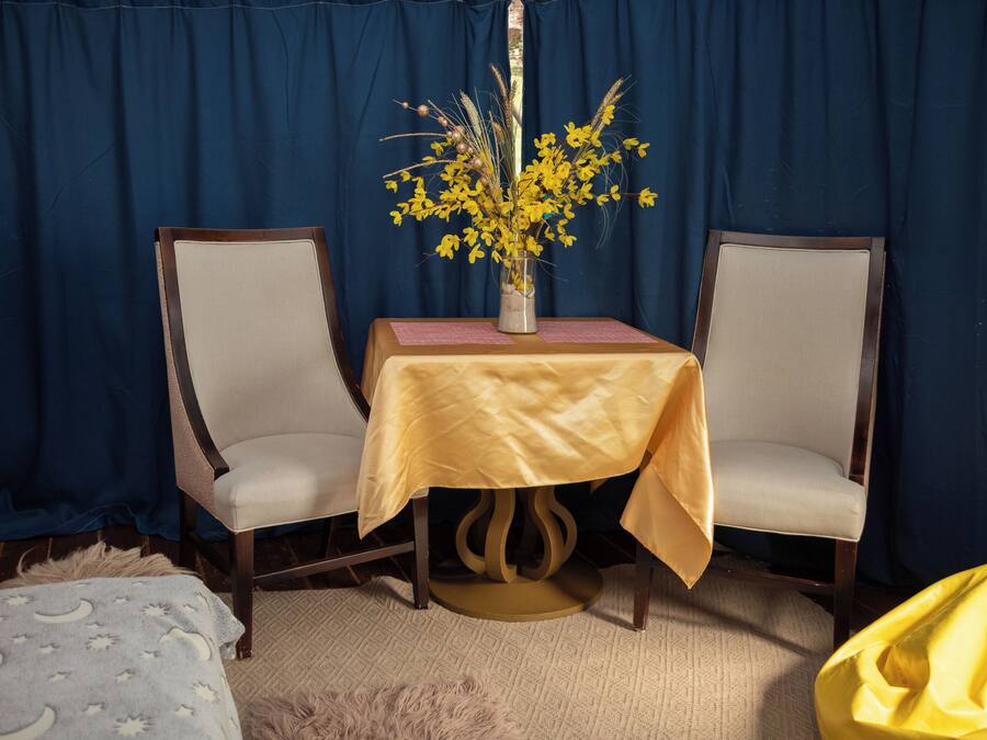 Una habitación con una mesa cubierta por un paño amarillo, dos sillas, un florero con flores amarillas y una cortina azul al fondo.
