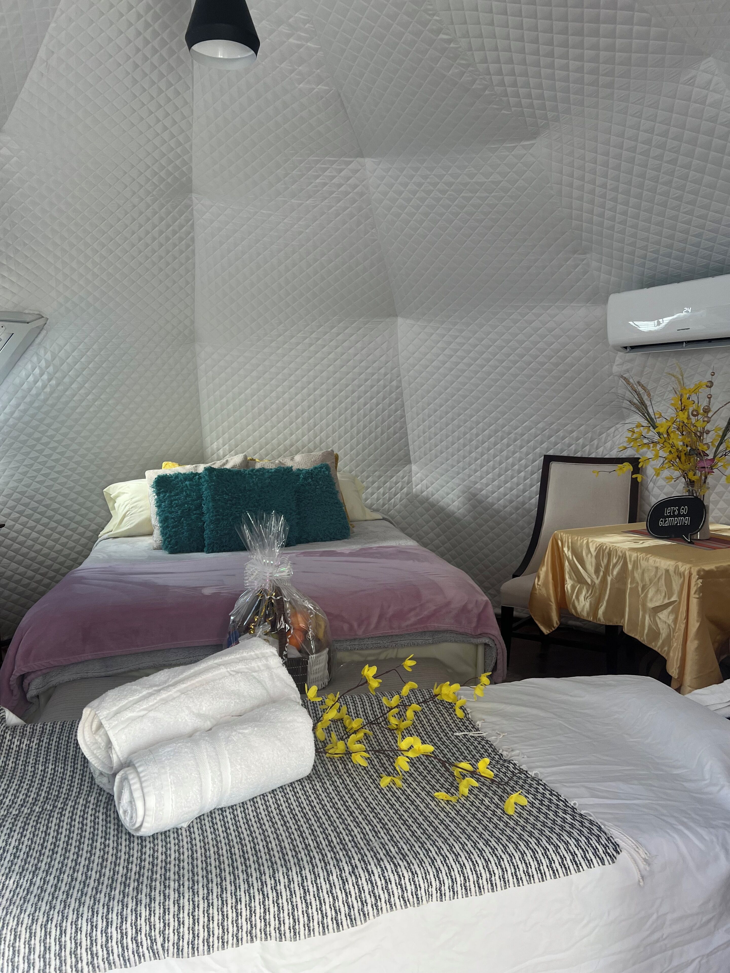 A room with a bed, a bedside table, a wall-mounted air conditioner, and a small table with a vase of flowers.