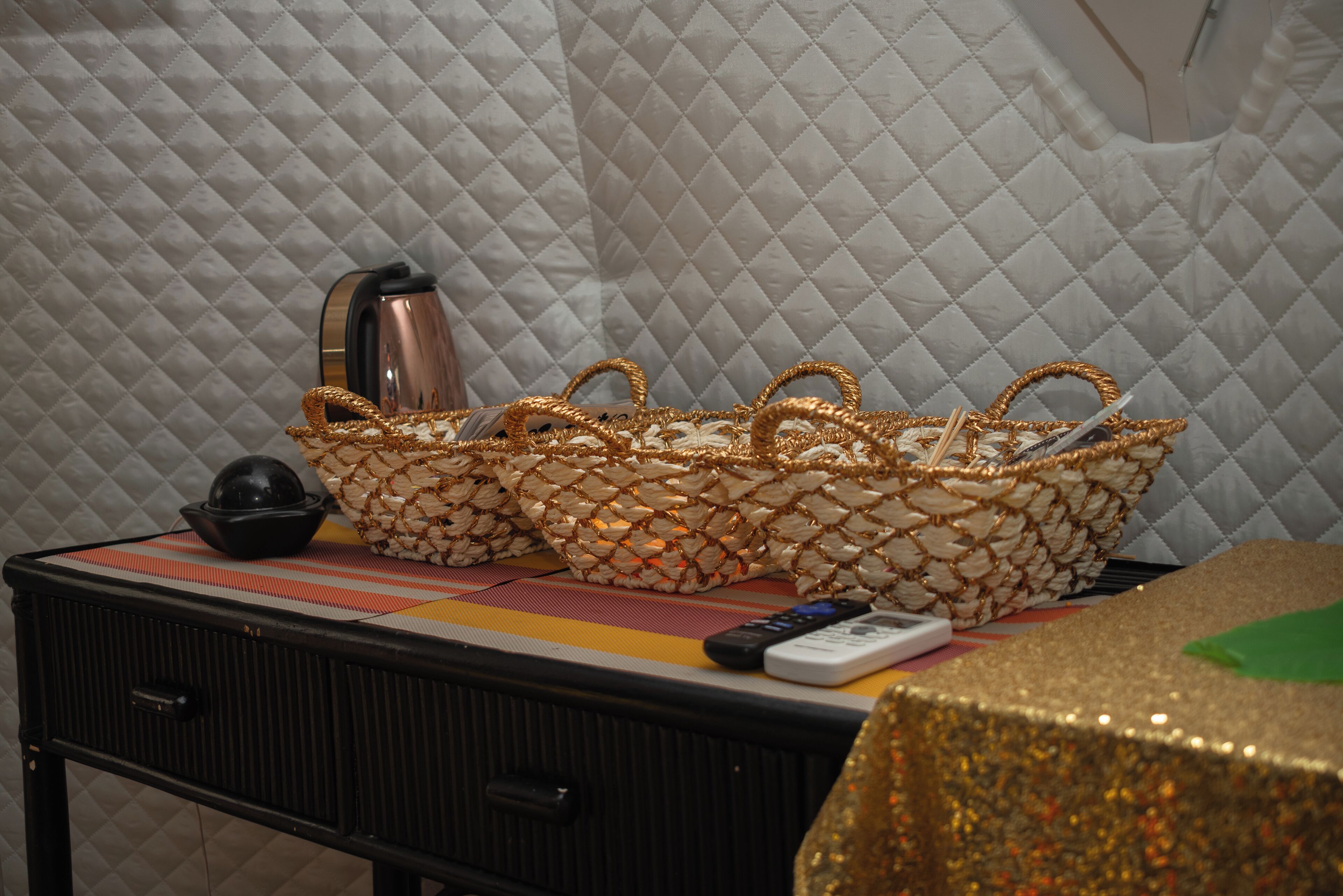 A bed with a quilted headboard, a bedside table with a remote control, a black lamp, and a decorative basket.