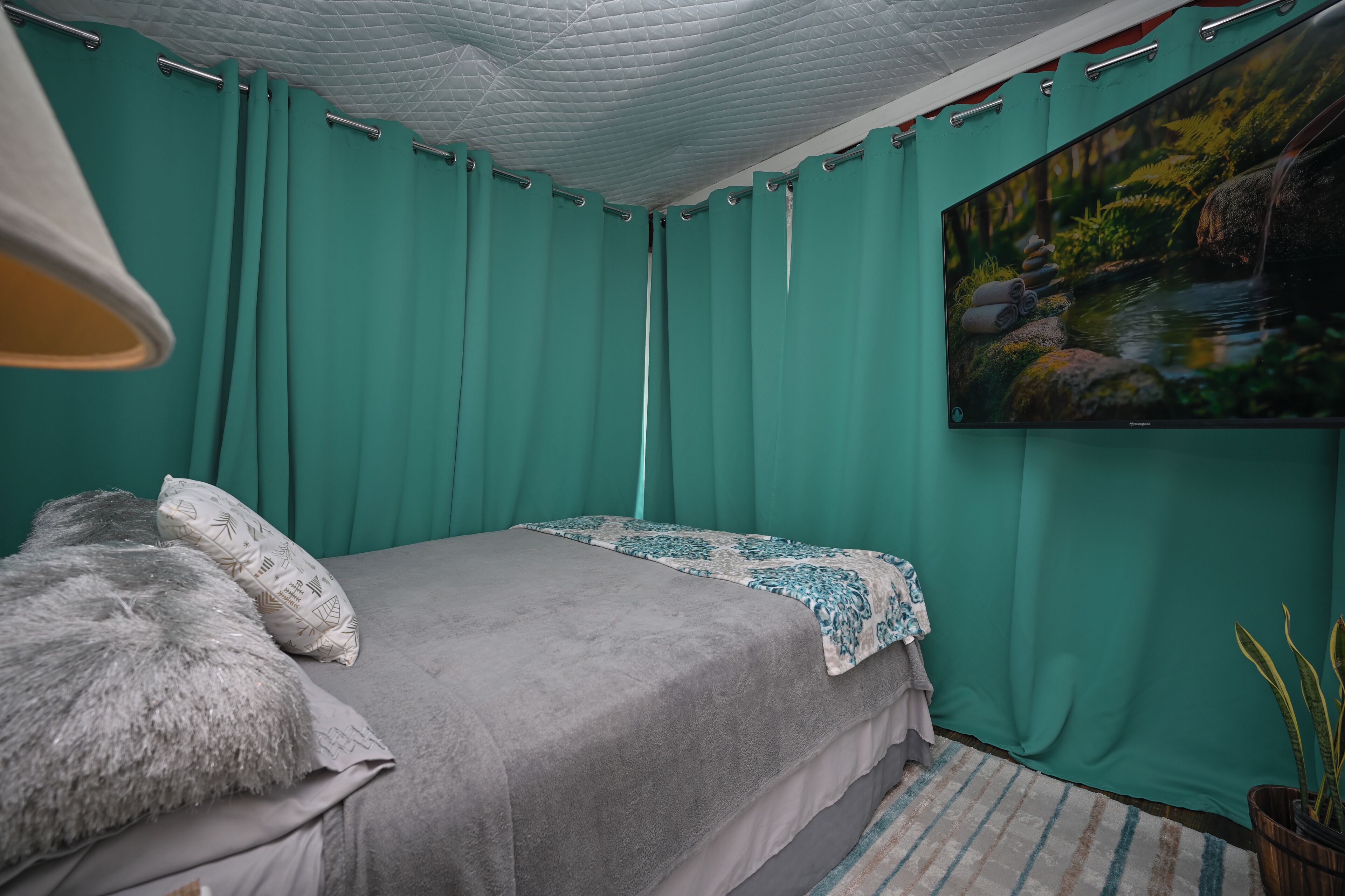 Une chambre avec des rideaux de couleur turquoise, un lit recouvert d’une couverture grise, un plaid à motifs, un tableau représentant un ruisseau de forêt et une plante en pot.