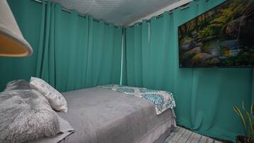 Une chambre avec des rideaux de couleur turquoise, un lit recouvert d’une couverture grise, un plaid à motifs, un tableau représentant un ruisseau de forêt et une plante en pot.