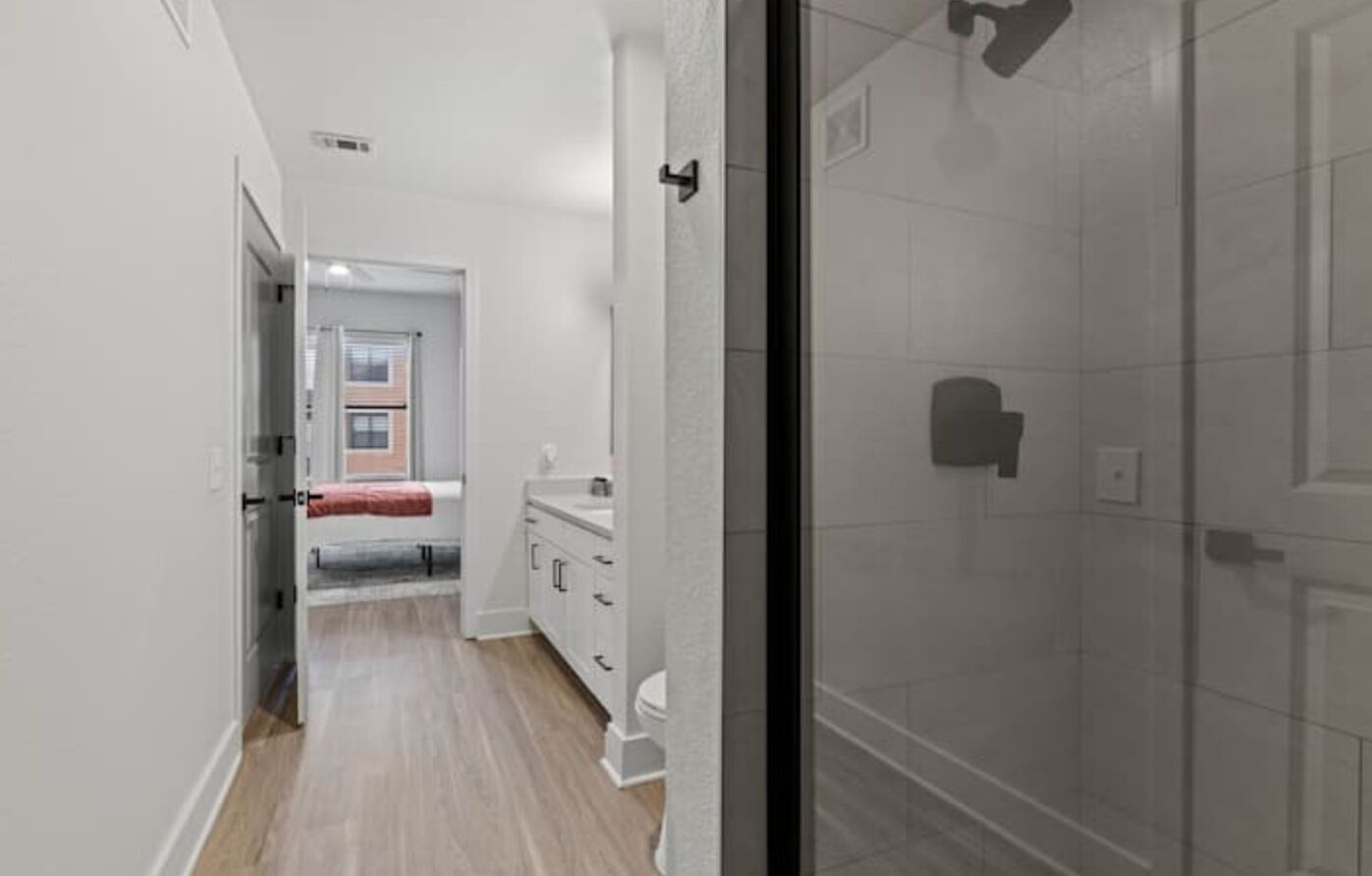 Un pasillo estrecho con un área de ducha a la derecha, una cama con colchón rojo al fondo y un baño blanco con lavamanos y espejo.