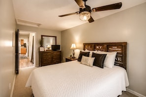 Un dormitorio con cama, ventilador de techo, cómoda, televisor y mesita de noche con lámpara.