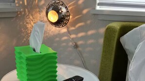 Hotelkamer met een groen tissuedoosje op een klein rond tafeltje, een afstandsbediening en een lamp met warm licht.