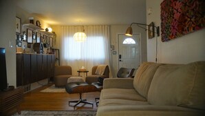 Un soggiorno con divano, poltrone, tavolino da caffè e televisore. Si nota una finestra con tende, una porta e una lampada a parete.