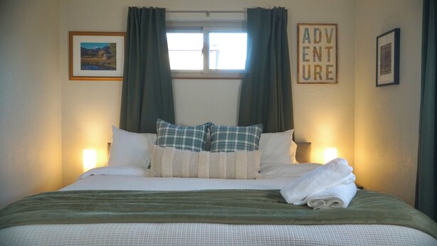 Un letto rifatto con cura, una coperta verde, due cuscini a quadri e un asciugamano piegato su un lato. Sono presenti due abat-jour, un quadro incorniciato appeso alla parete e un cartello con la scritta 'ADVENTURE'.
