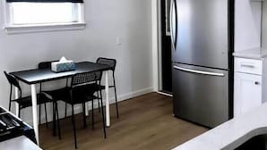 A compact kitchen with a stainless steel refrigerator, a dining table with chairs, and a window with blinds.