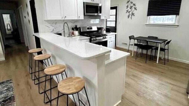 A modern kitchen with white cabinets, a central island with stools, a dining area with a table and chairs, and a window with blinds.