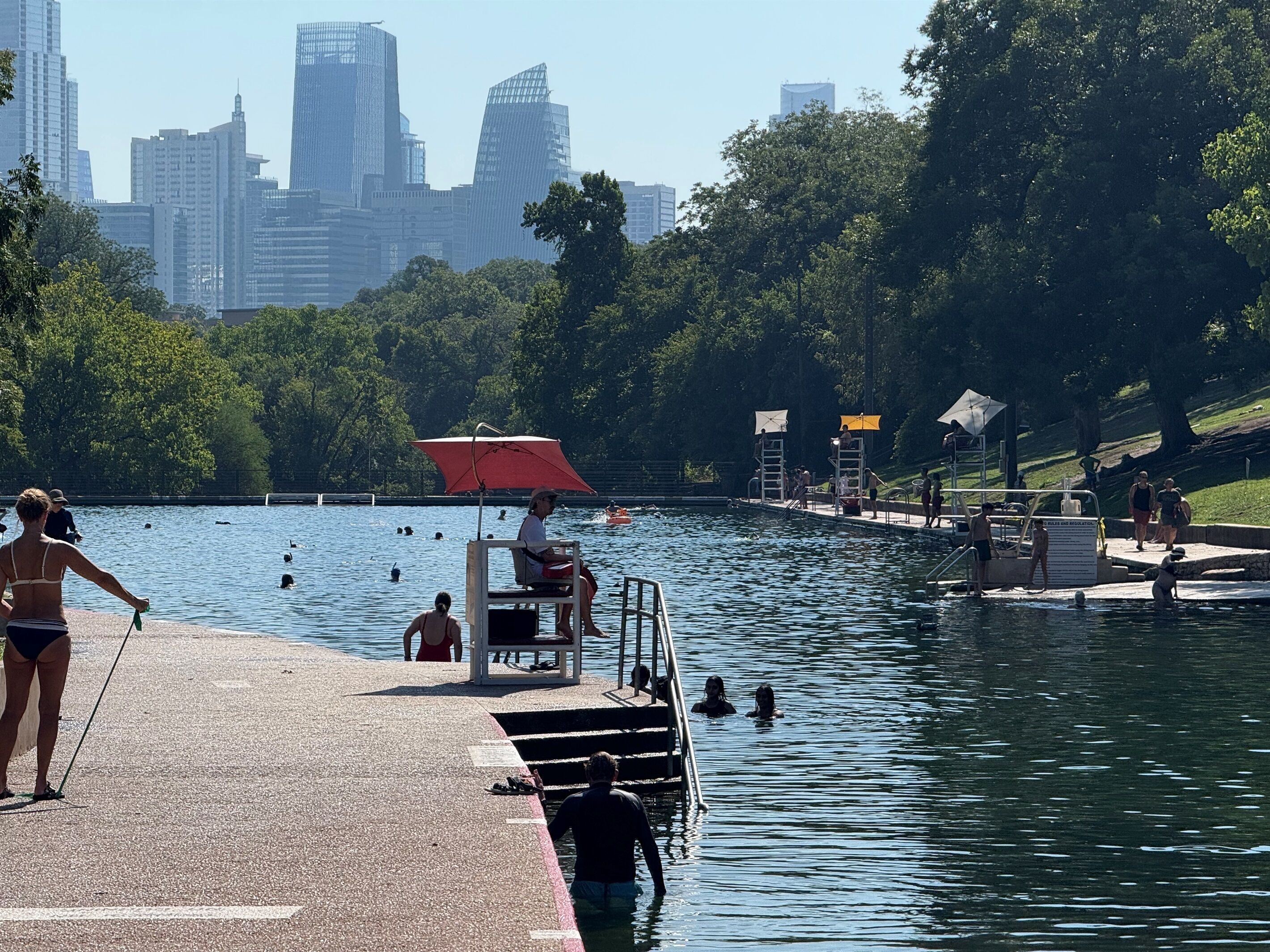 A cityscape with modern buildings in the background, a swimming pool area with people, and a dock with a lifeguard chair and umbrella.