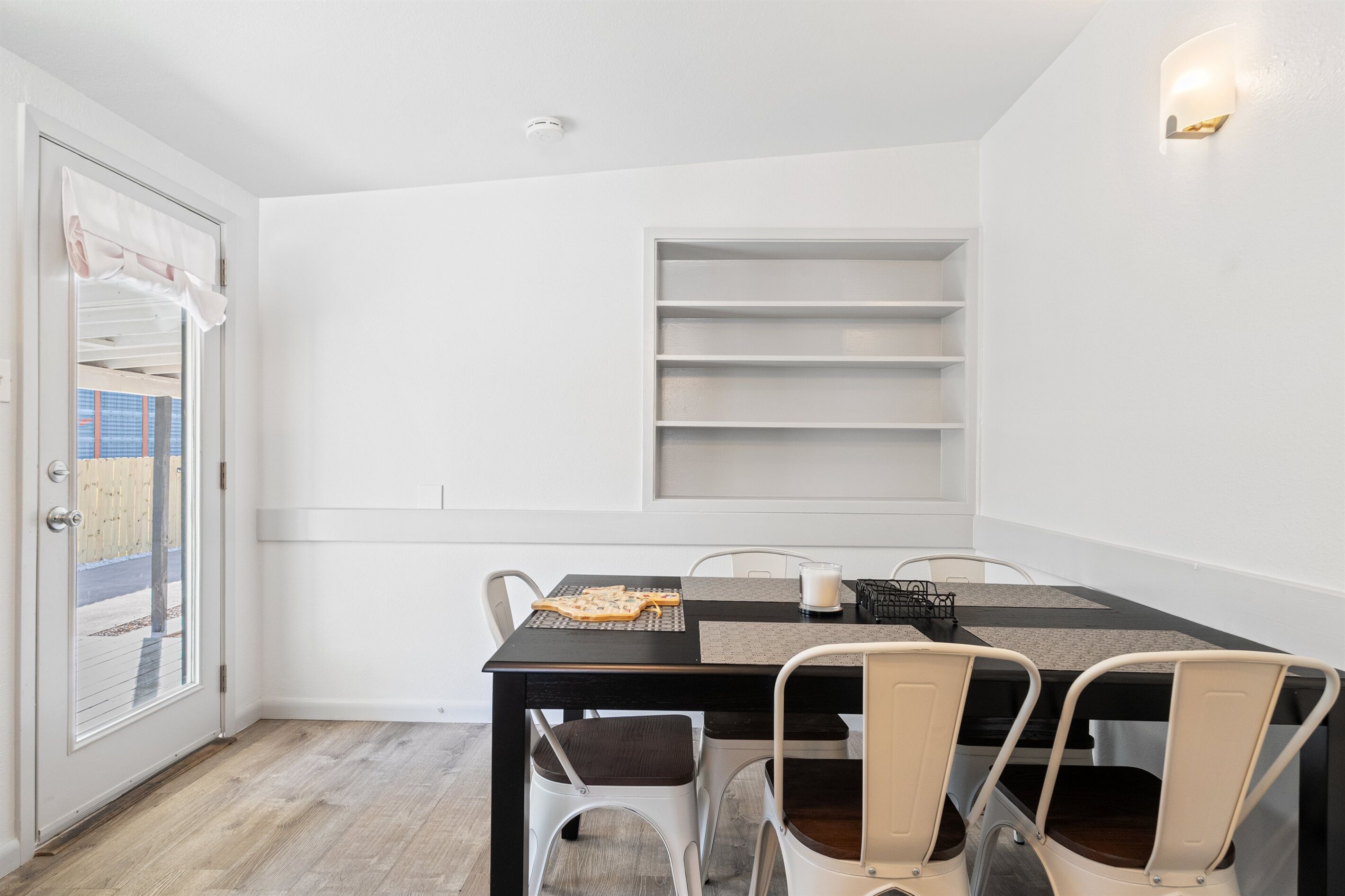 A dining area with a black table, white chairs, and a white door with a view of the outside.