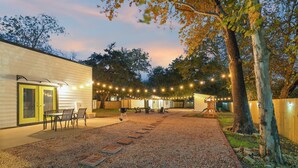A covered outdoor patio with string lights, a barbecue grill, and a gravel pathway leading to a building with a door and windows. - Austin Gated Oasis | 4BR | Sleeps 8 | Fire Pit (Austin)