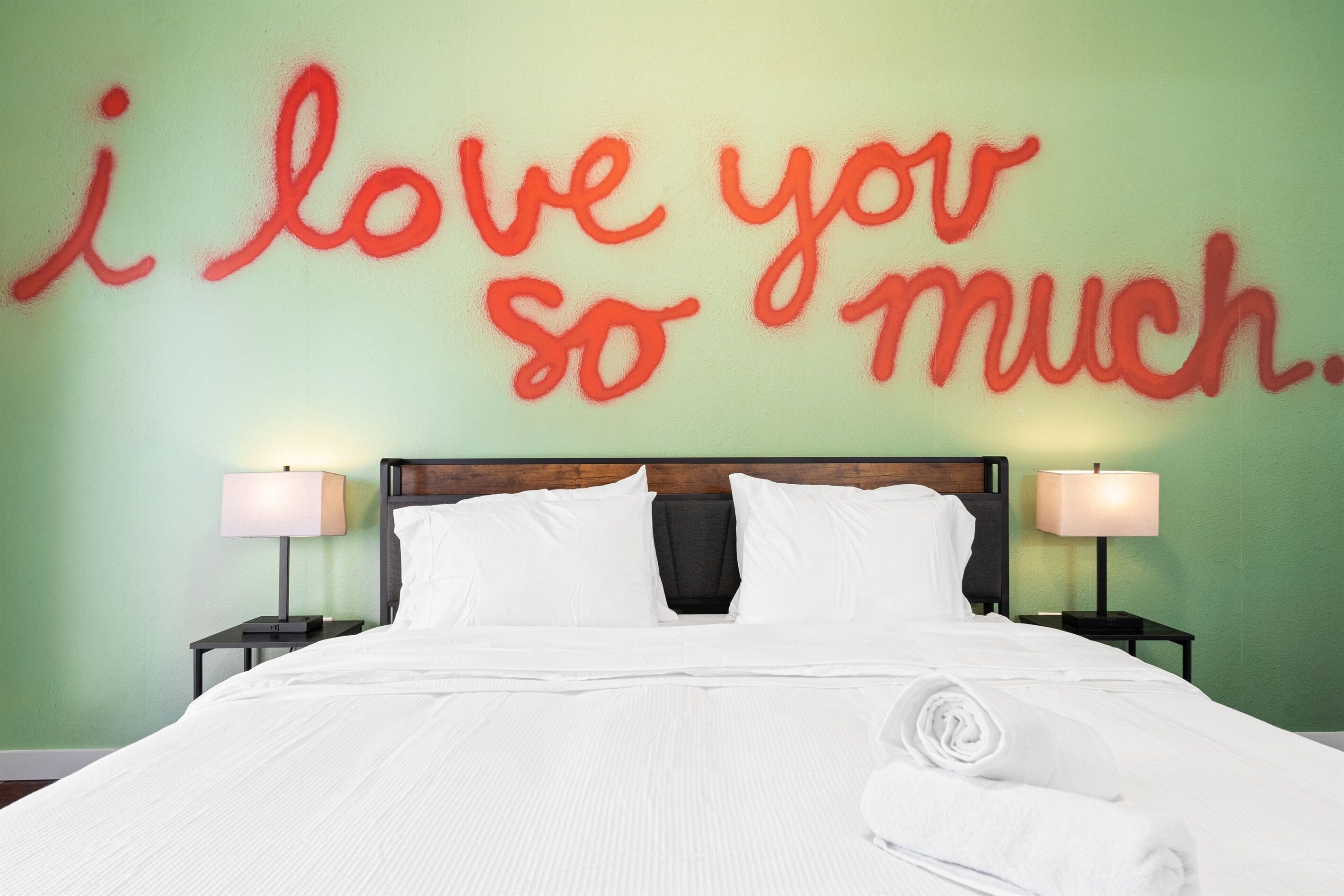 A neatly made bed with white linens and a rolled towel, a wooden headboard, two bedside lamps, and a green wall with a red phrase saying "I love you so much."