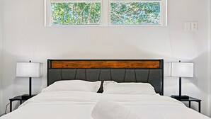 A neatly made bed with white linens and a wooden headboard, flanked by two bedside tables with lamps, in a room with white walls and a window with a view of trees. - Austin Gated Oasis | 4BR | Sleeps 8 | Fire Pit (Austin)