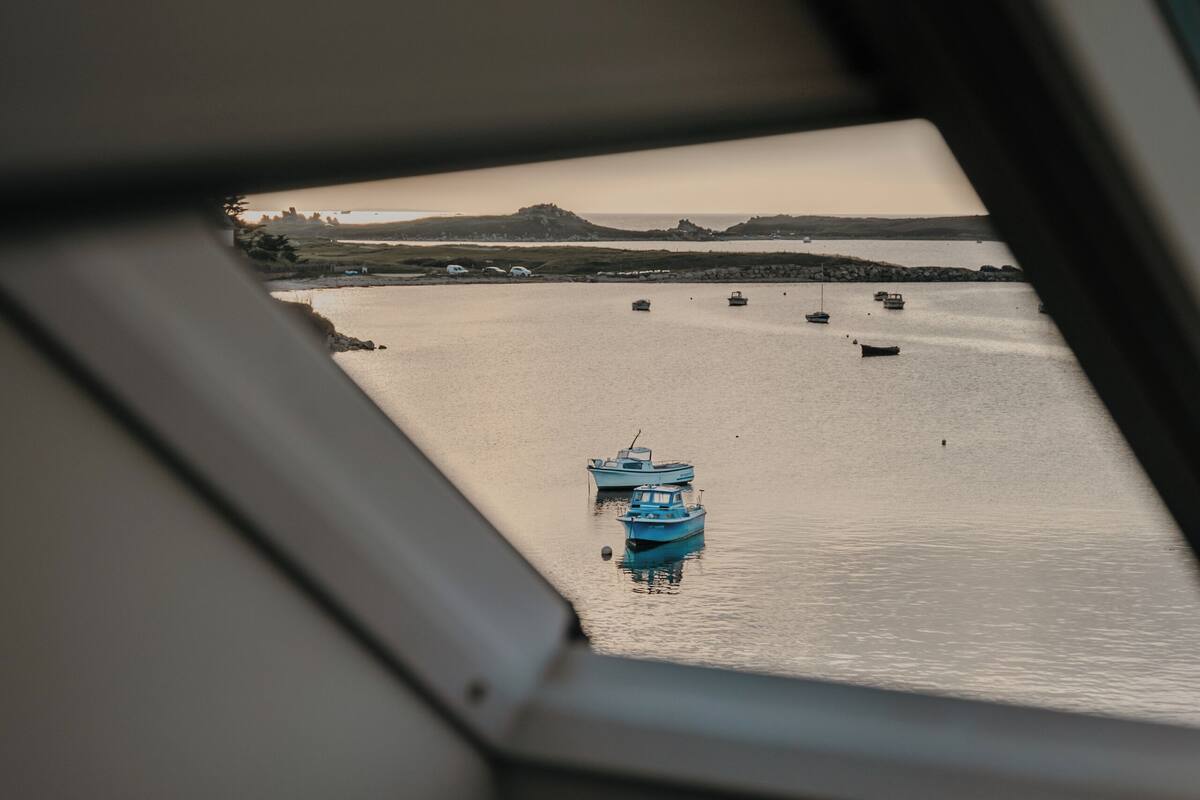 Μια θέα μέσα από ένα παράθυρο: μια ήρεμη επιφάνεια νερού με αρκετές βάρκες, μια μακρινή ακτογραμμή και ένας συννεφιασμένος ουρανός.