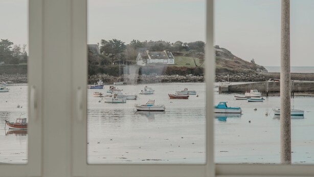 Blick durch ein Fenster auf eine Küstenlandschaft mit Booten und einer Hügelkette im Hintergrund.