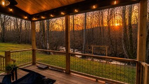 A wooden deck with a railing, benches, and a view of a forest during sunset. - Saddle Tree - Etowah River - Firepit - New Build (Dahlonega)