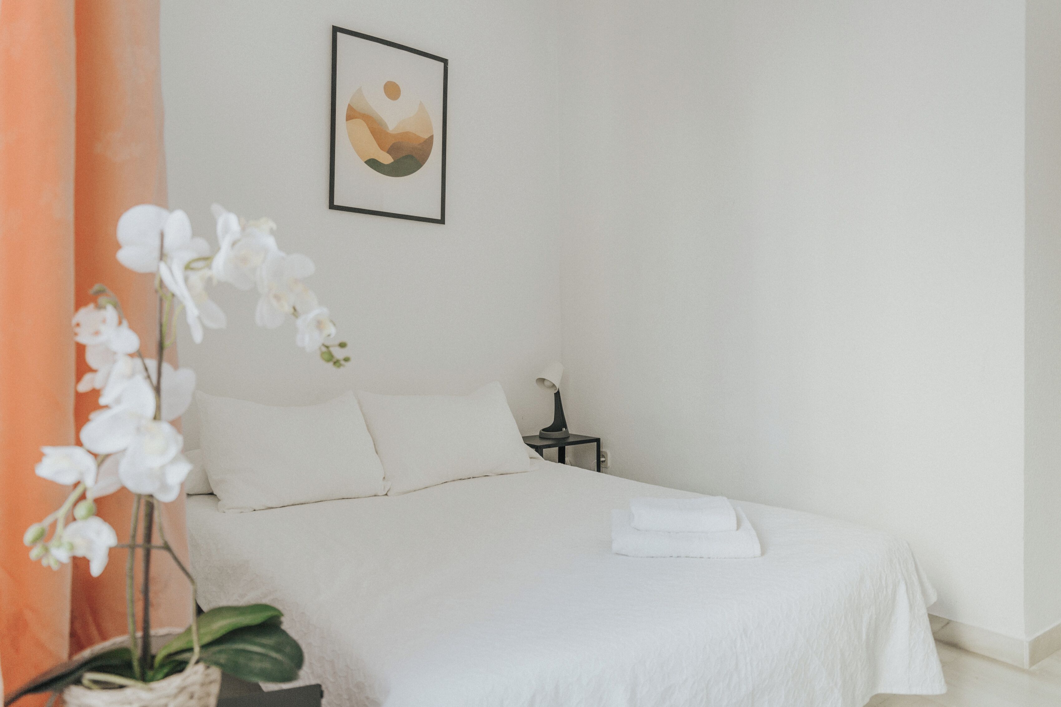 A neatly made bed with white linens, a small bedside table with a lamp, and a framed picture on the wall.