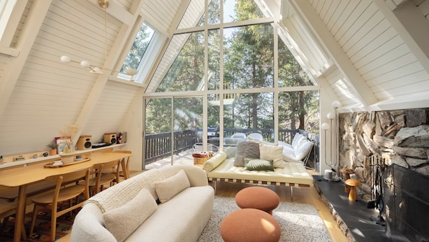 A cozy, well-lit room with a high ceiling, a fireplace, a dining area, and a large window offering a view of the outdoors. - A-Frame + Indoor Sauna & Hot Tub · Lake Arrowhead (Lake Arrowhead)