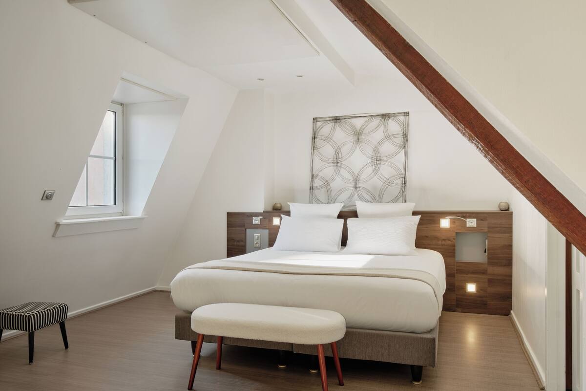 A modern bedroom with a sloped ceiling, a large bed, a bench, and a window.