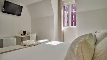 A hotel room with a bed, a flat-screen TV mounted on the wall, a small table with a lamp, and a window with a view of a building.