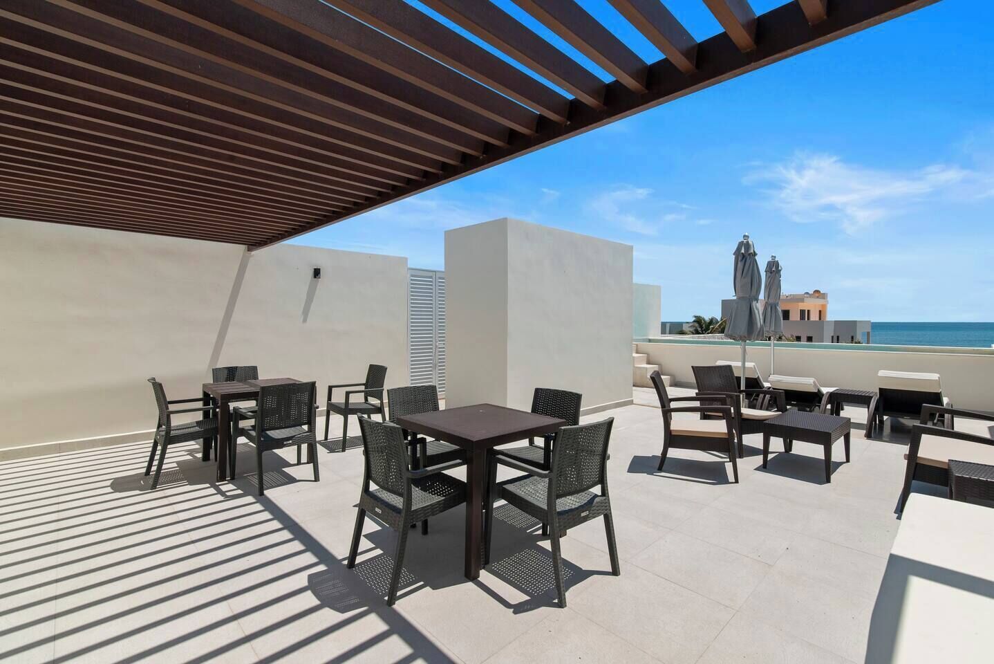 An outdoor patio with tables and chairs, a pergola, and a view of the sea and buildings in the background.