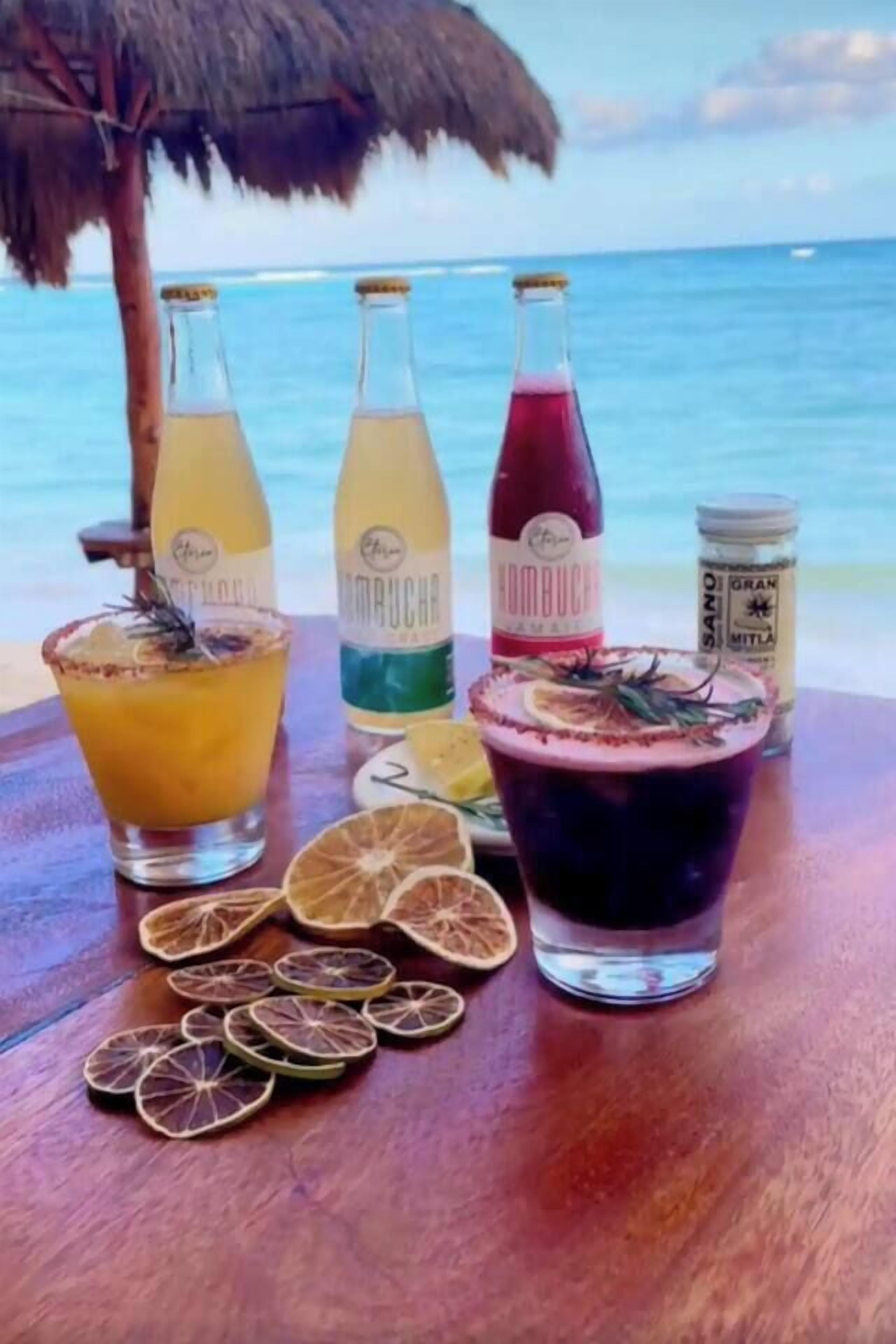 A table with three bottles of kombucha and a large glass of a dark beverage, surrounded by dried citrus slices.
