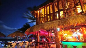 Restaurante à beira-mar com mesas ao ar livre, guarda-sóis de palha e um bar.
